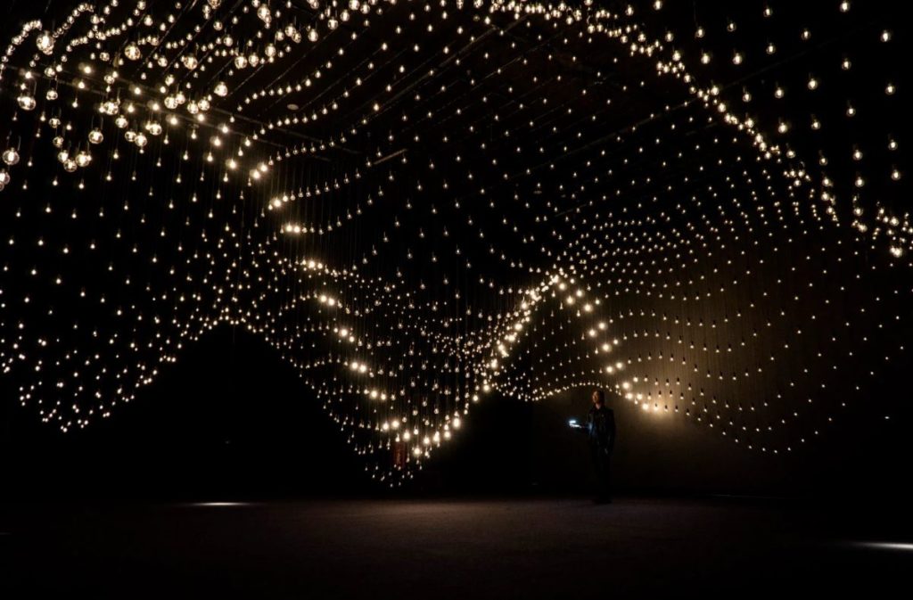 A person stands beneath hundreds of suspended lightbulbs in a flowing pattern.