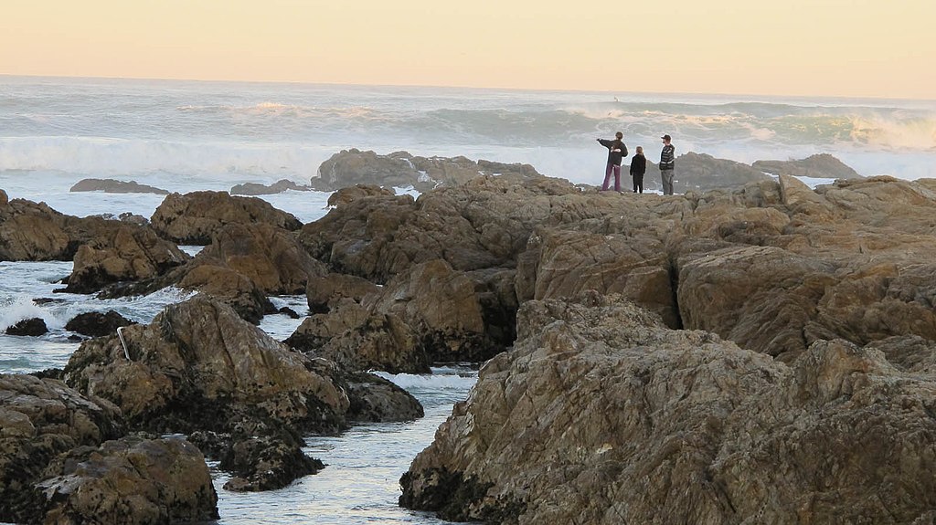Three people stand on the rocks at sunset at Asilomar State Beach