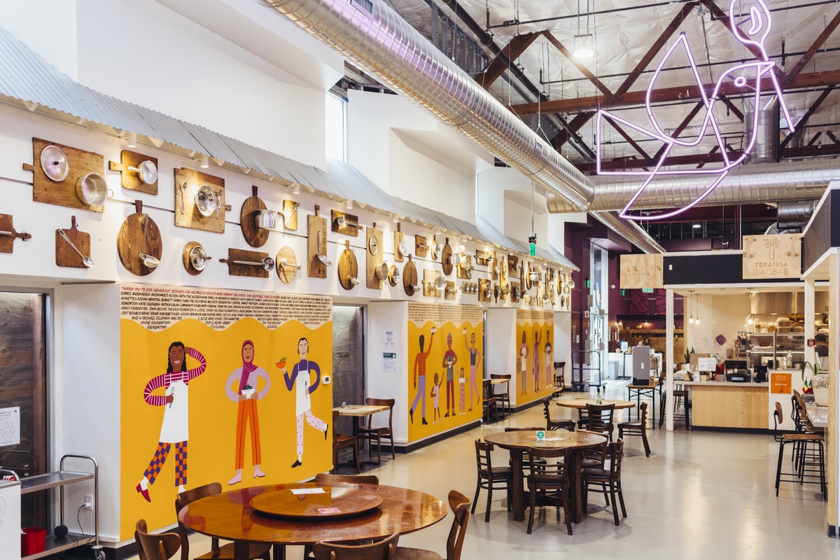 Interior of La Cocina Municipal Marketplace, depicting tables and chairs before a yellow mural thanking donors.
