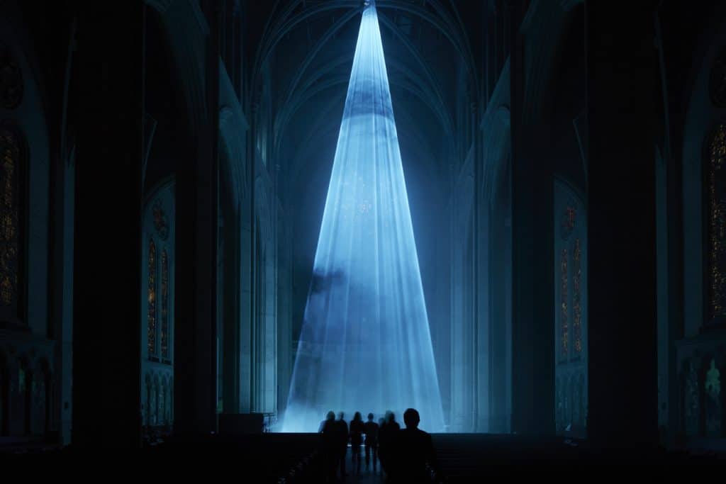 A tall luminous light blue beam of light shines down on a silhouetted group of people in a dark cathedral.