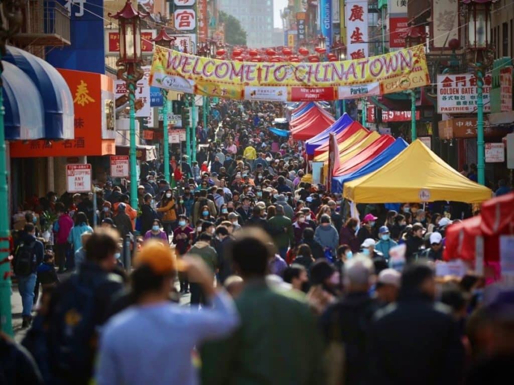 A massive crowd of people takes up a colorful street with a banner over it that reads 