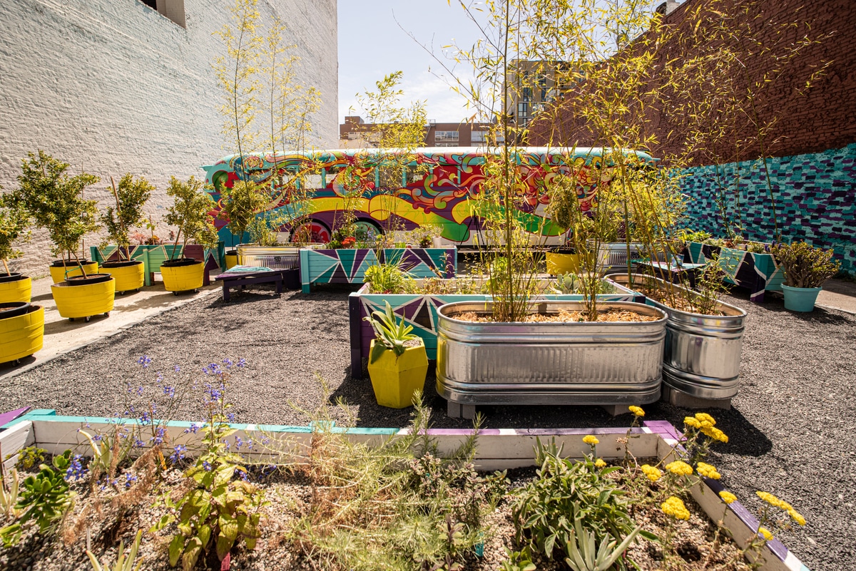 Kapwa Gardens' garden space, with large metal planters and leafy plants in front of their colorful bus backdrop.