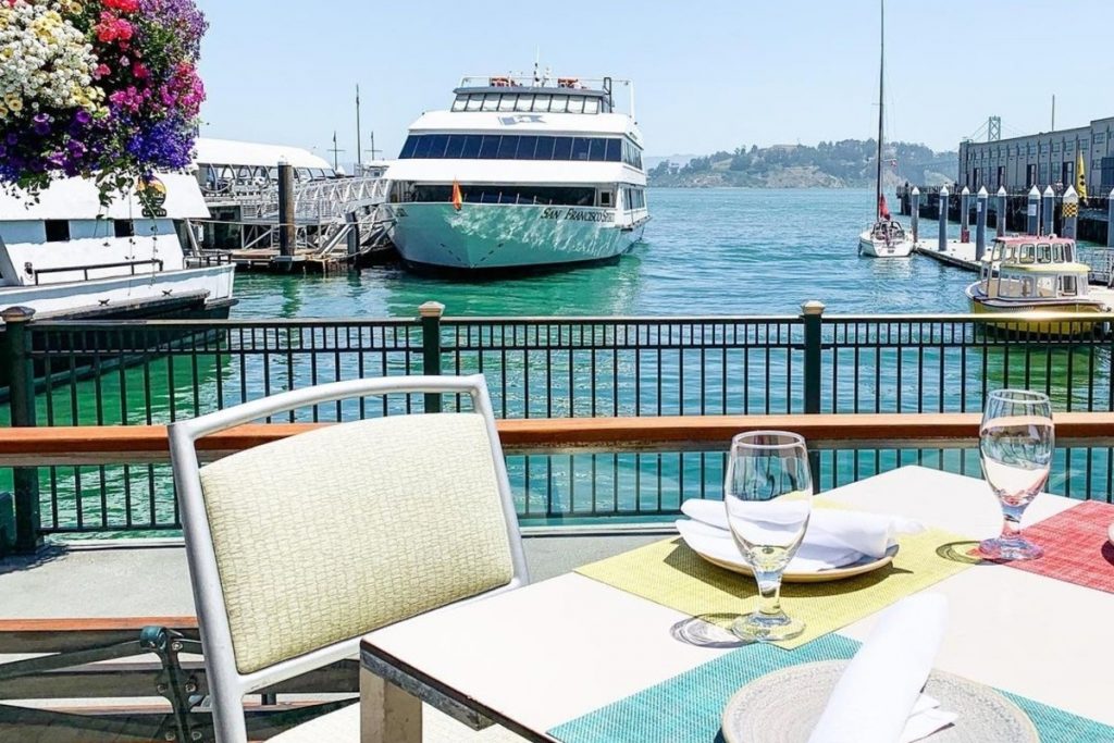 An outdoor table set up with plates and wine glasses on the waterfront with a view of the SF Bay and ships in the background.