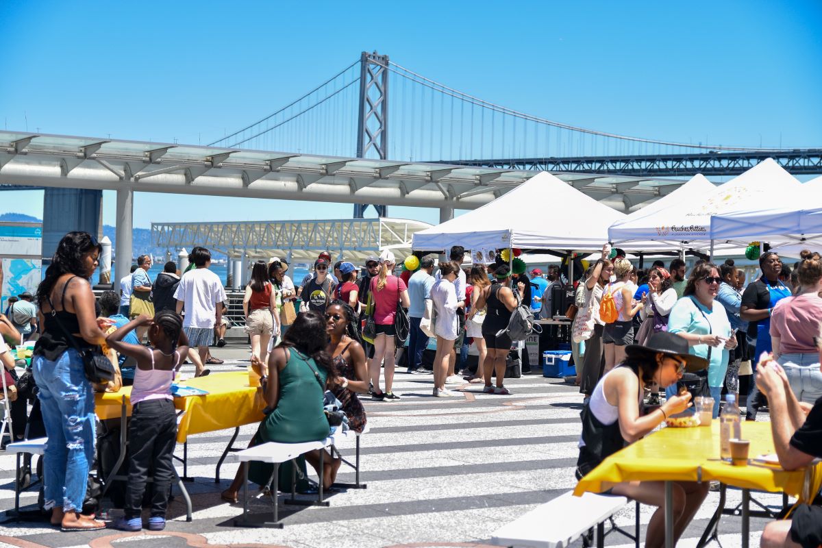 Groups of people walk around vendors stations and tents on the Ferry Building plaza with the Bay Bridge in the background.