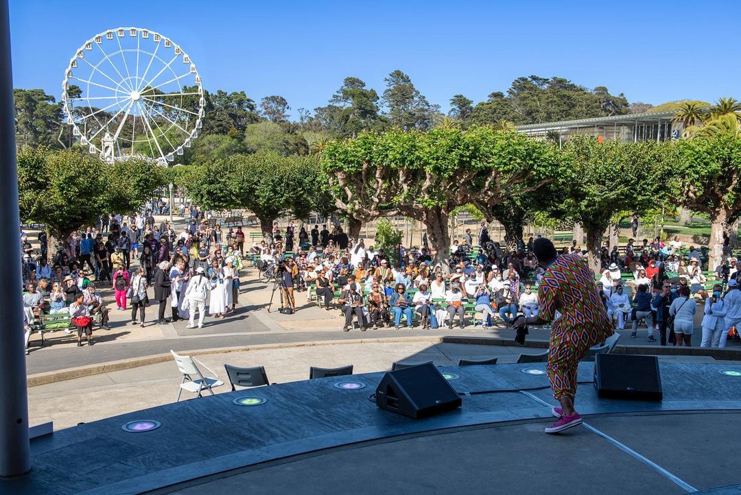 A singer performs in front of a crowd at Golden Gate Park's Bandshell. The picture is from behind the singer looking out at the Music Concourse's green trees and SkyStar Ferris wheel.