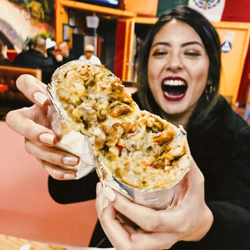 A smiling woman holds a burrito up to the camera.