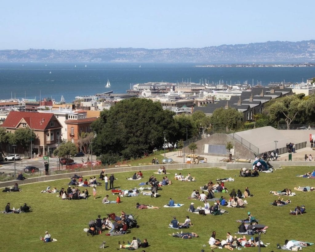 Groups of people picnic on the lawn at Francisco Park, with a view of the ocean in the background.