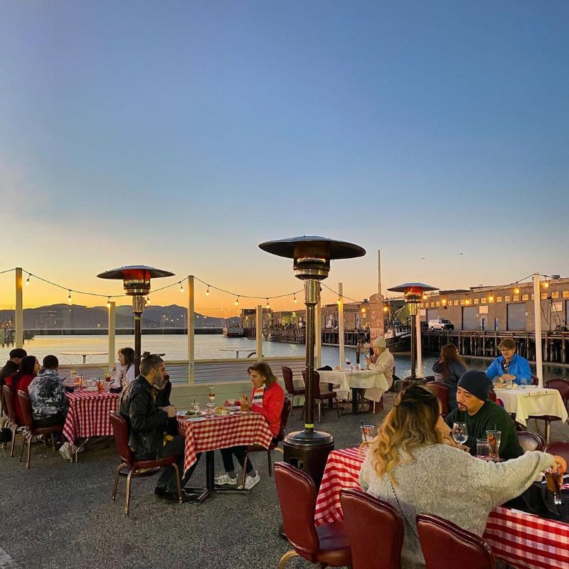Diners eat at Scoma's outdoor patio with red checkered tablecloths and a sunset view.