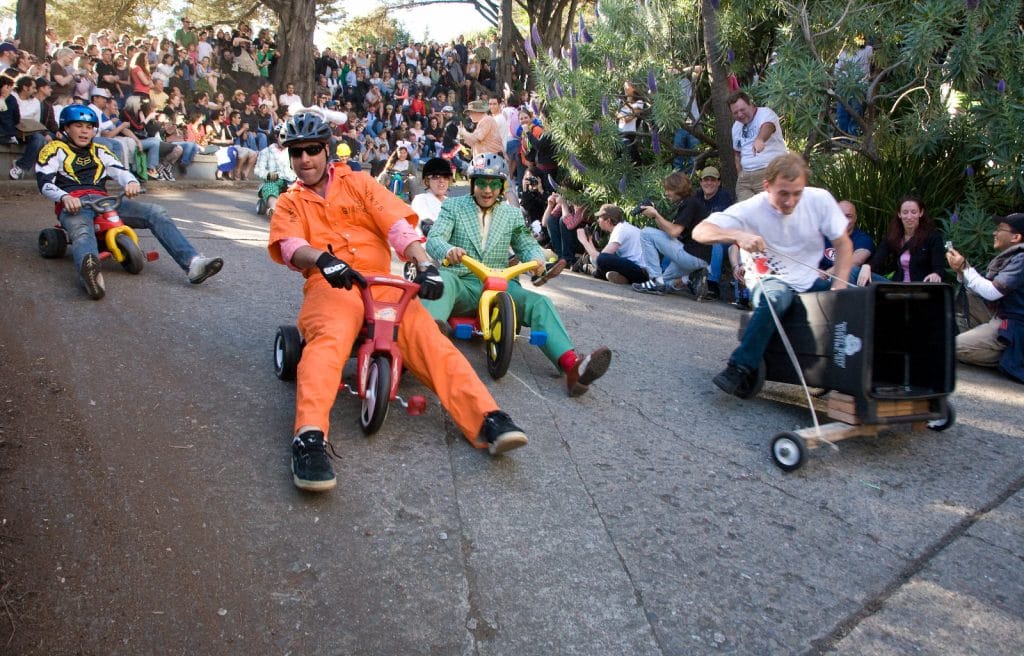 A group of people in costumes race down a hill on big wheel bikes.