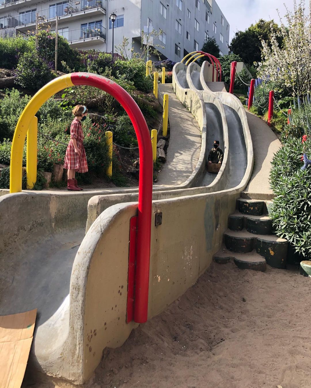 Two large concrete slides with red and blue accents wind down a hill. A little girl in a red plaid dress watches as another child comes down the slide.
