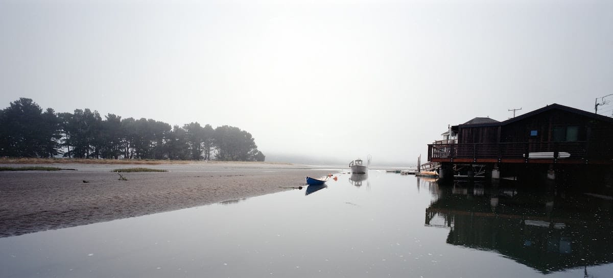 A foggy day at the beach with several boats docked on smooth water near a dark wooden building.