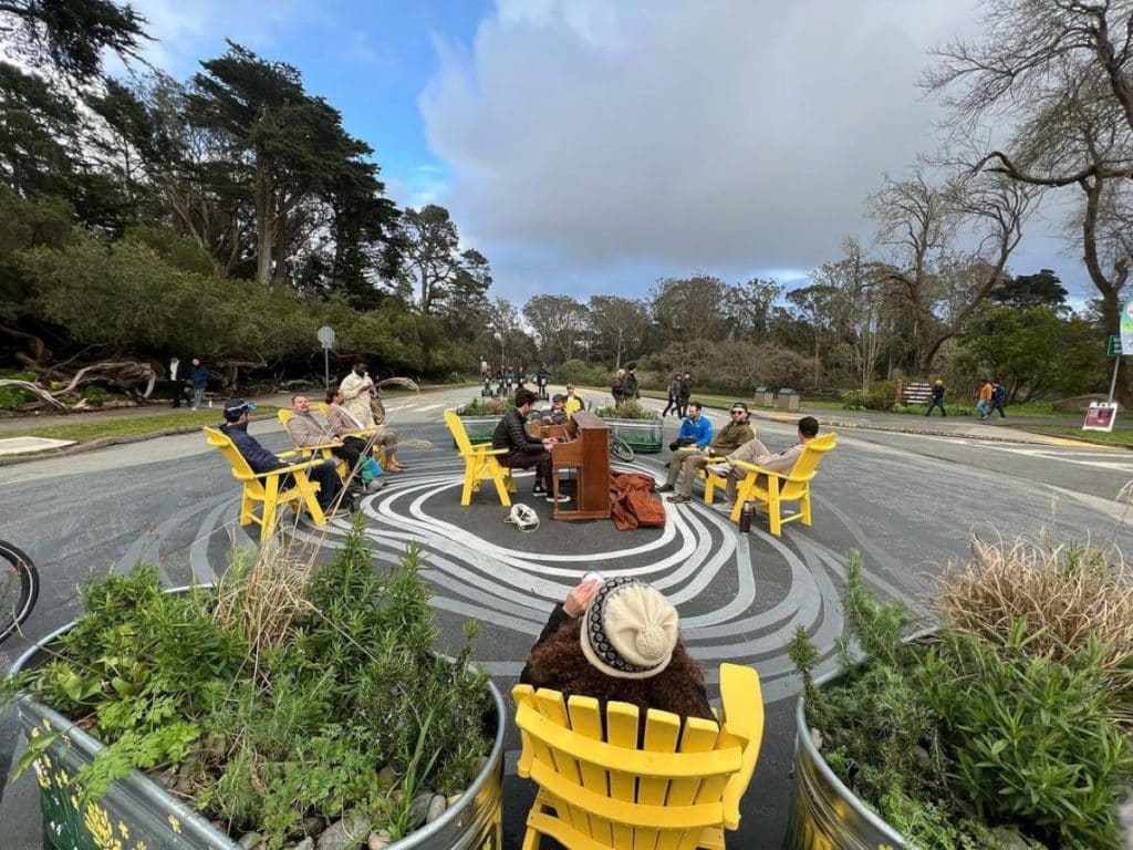 People sit in yellow Adirondack chairs watching someone perform on a public piano surrounded by a ground mural on JFK Drive.