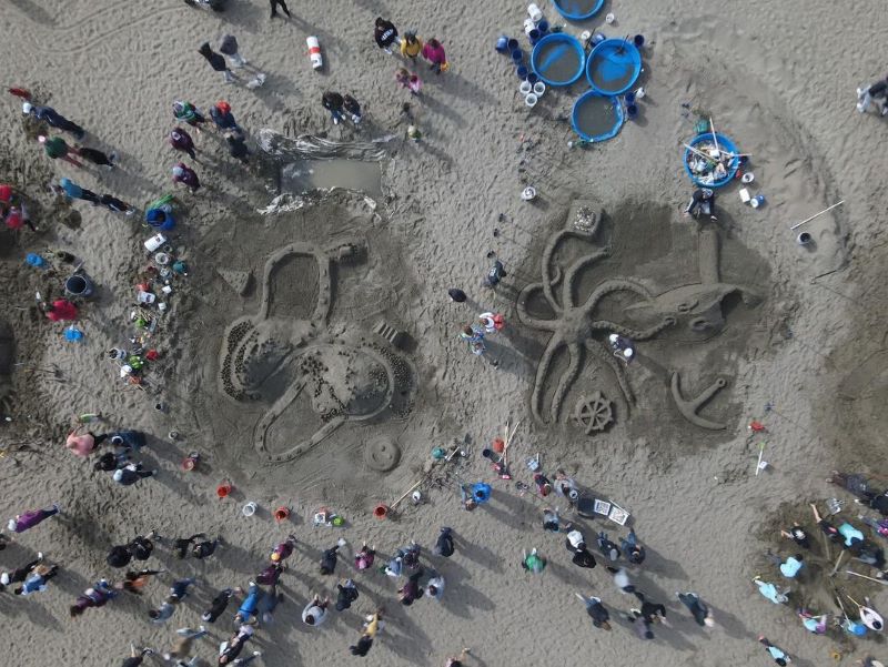 Drone shot from above of groups of people working on two giant sandcastles side-by-side. 