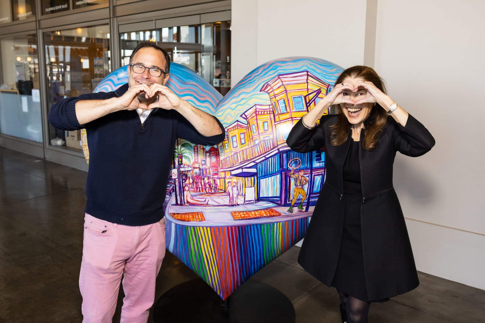 A man and a woman pose making heart shapes with their hands in front of a heart sculpture depicting a colorful SF city street. 