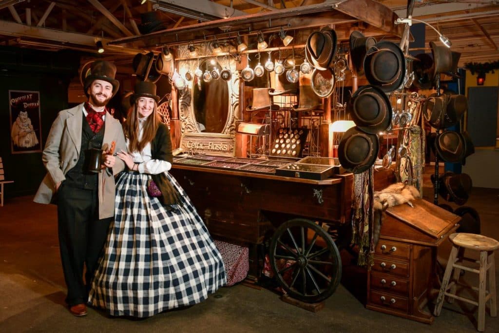 A couple in Victorian garb poses next to an antique stand selling Celtic flasks at the Dickens Fair.