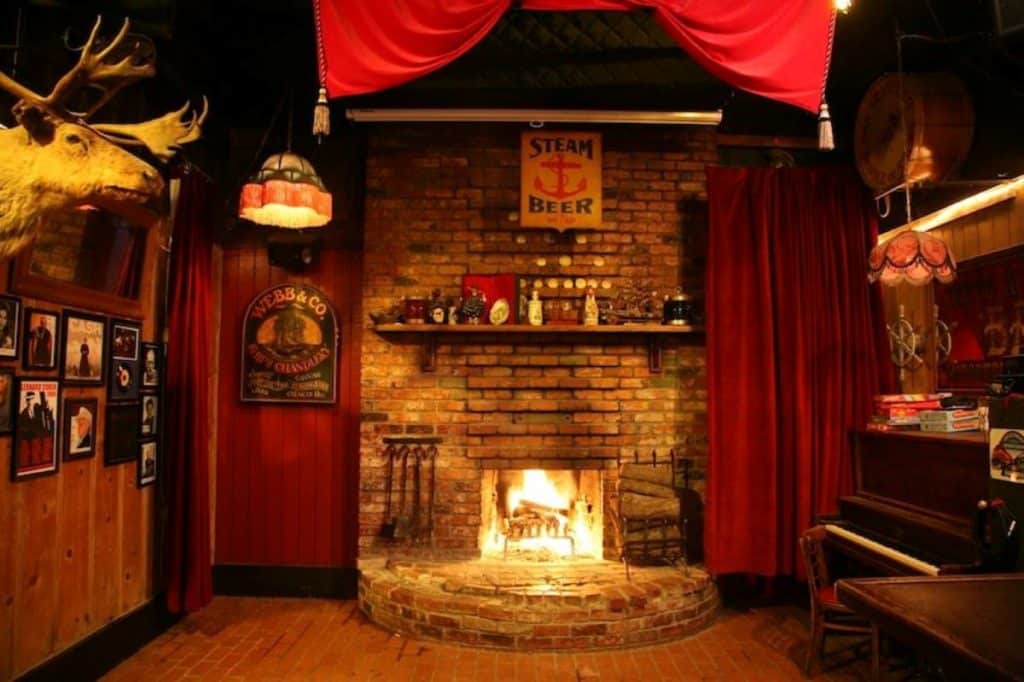 A stone fireplace with a big fire in a room with red curtains and a brick wall.