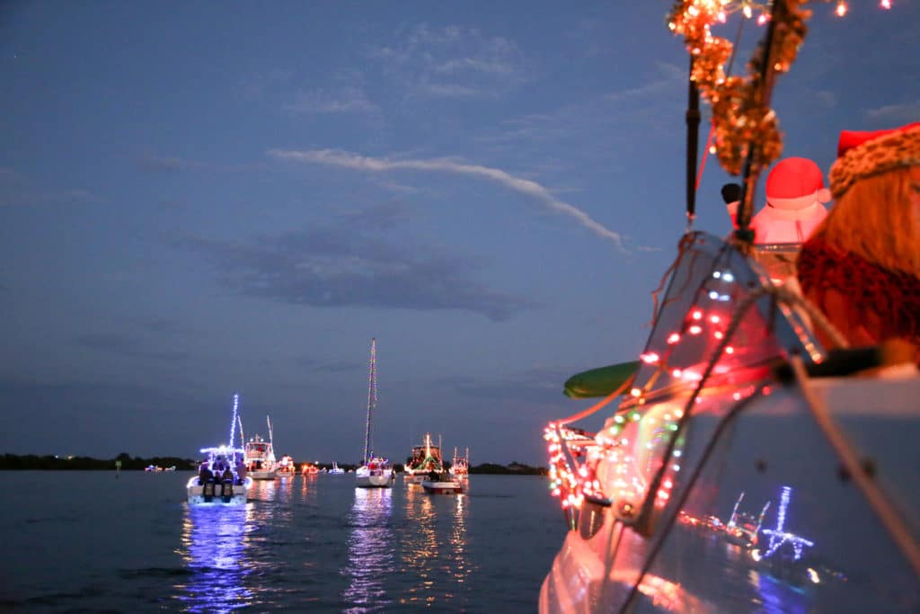 A group of sailboats decorated with holiday lights takes to the water at dusk.