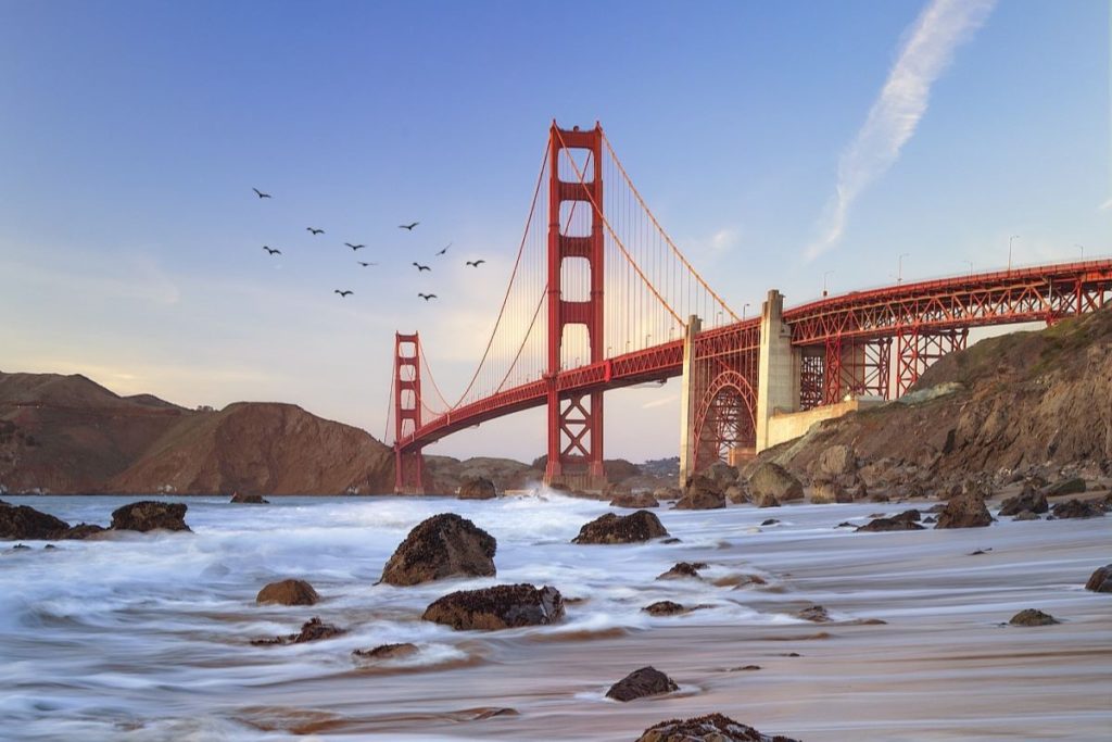 Golden Gate Bridge with beach in foreground and flock of birds flying in background.