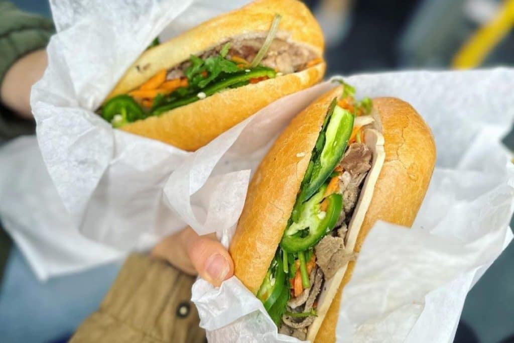 A close-up of two banh mi sandwiches with green peppers, carrots, and a cooked meat.