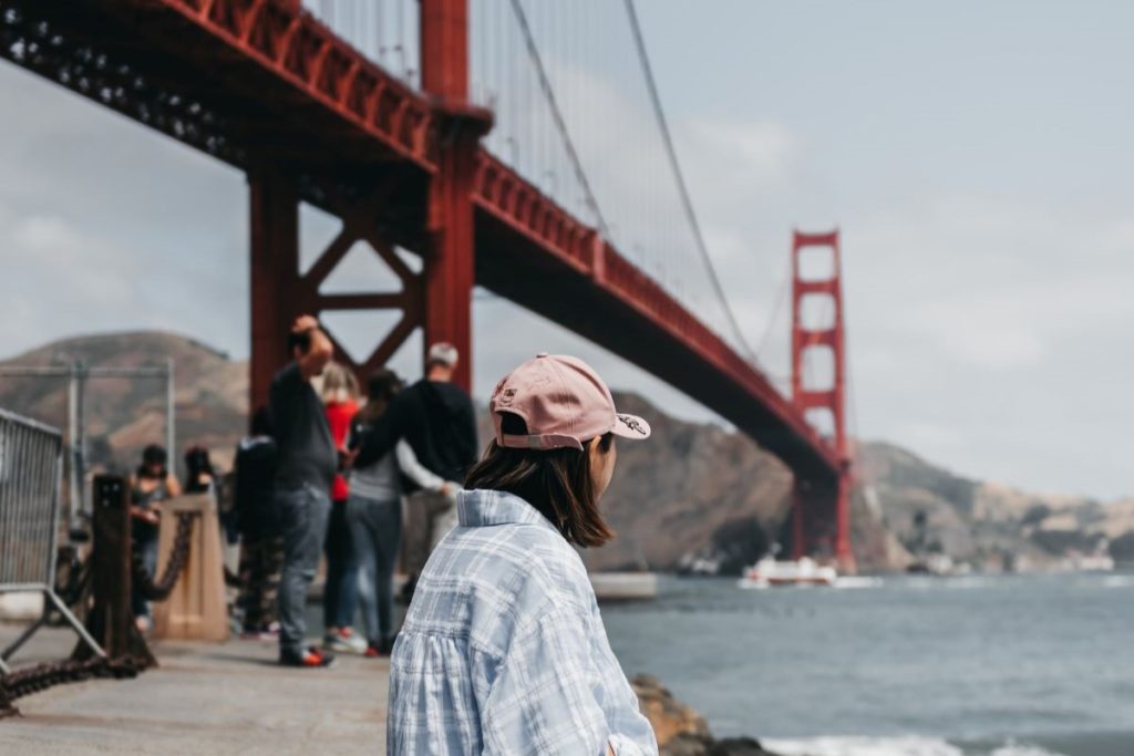 A seated woman in a pink hat faces away from the camera looking at the Golden Gate Bridge and SF Bay.