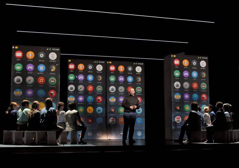 An actor portraying Steve Jobs stands on a stage surrounded by recurring images of a smartphone home screen. 