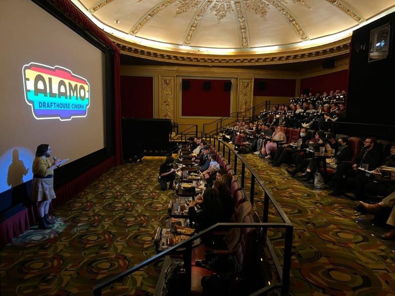 A crowd of people in an ornate room with stadium seating watch a speaker with a microphone in front of a screen reading 'Alamo Drafthouse Cinema'