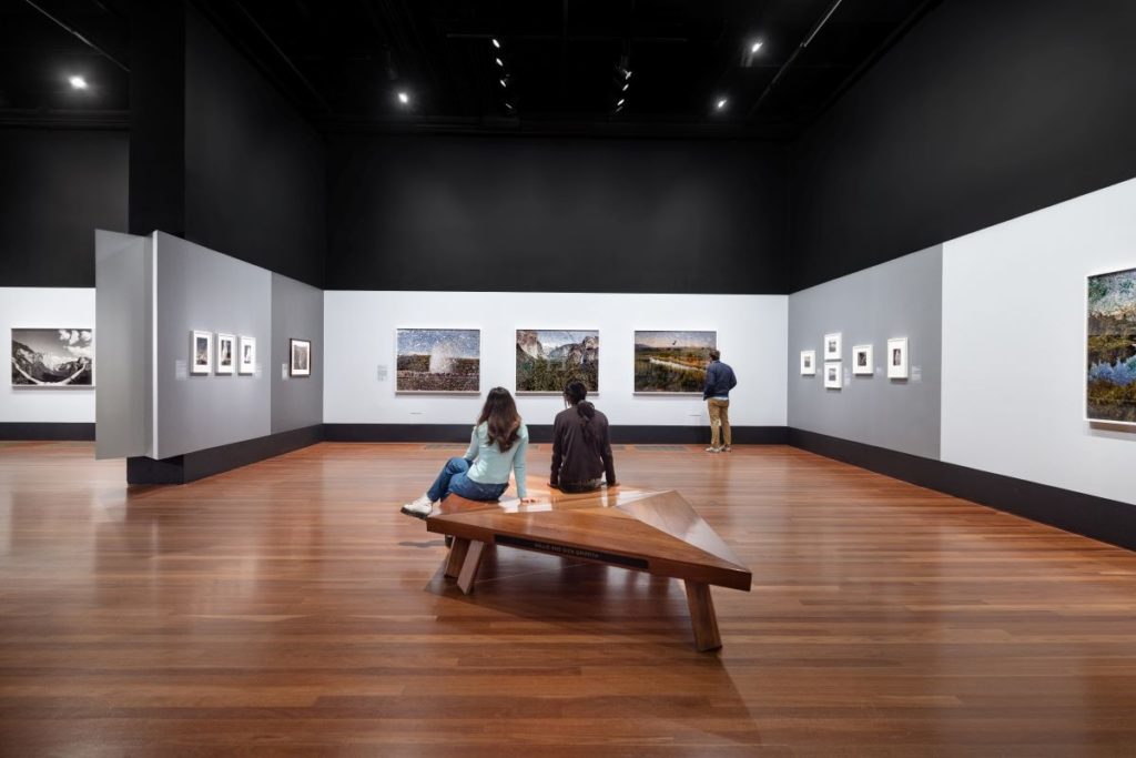 Two people sit on a diamond-shaped wooden bench at the Ansel Adams exhibition.