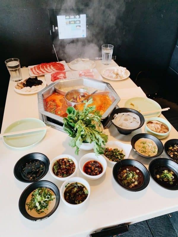 A hot pot table with a steaming bowl of soup in the middle surrounded by sides.