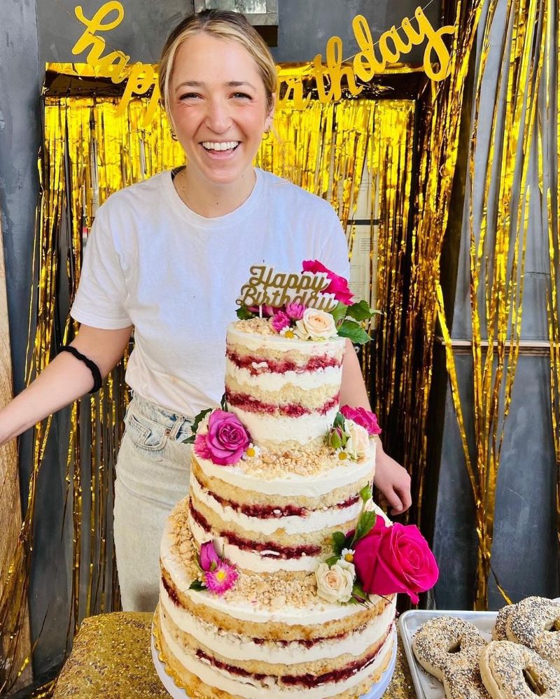 Butter and Crumble owner Sophie Smith smiles next to a 3-tier cake with the words "Happy Birthday."