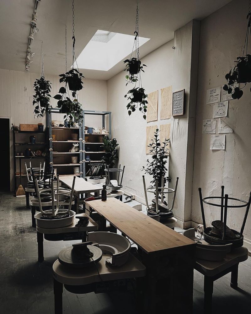 A small empty pottery studio with a line of pottery wheels and hanging plants.