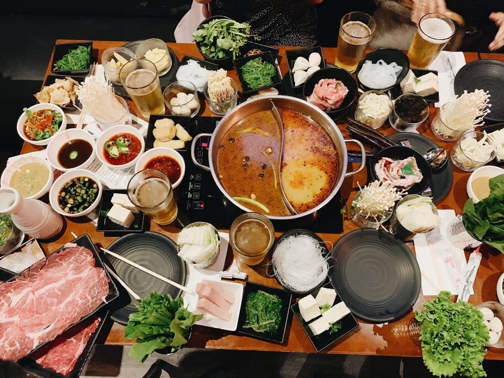 A hot pot table with a steaming pot of soup in the center surrounded by dozens of small plates of assorted meats and veggies.