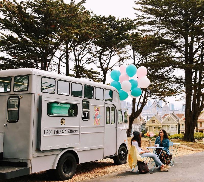 Two women drink coffee at a table outside next to a gray converted bus with the words "Lady Falcon Coffee Club" on the side, decorated with blue and white balloons.