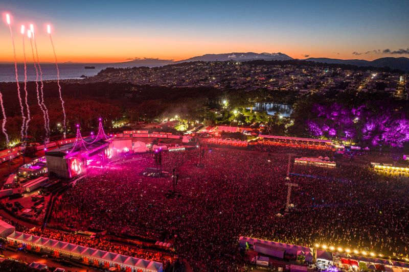 A birds-eye view of Outside Lands with thousands of people packed into the festival grounds and a sunset in the background.