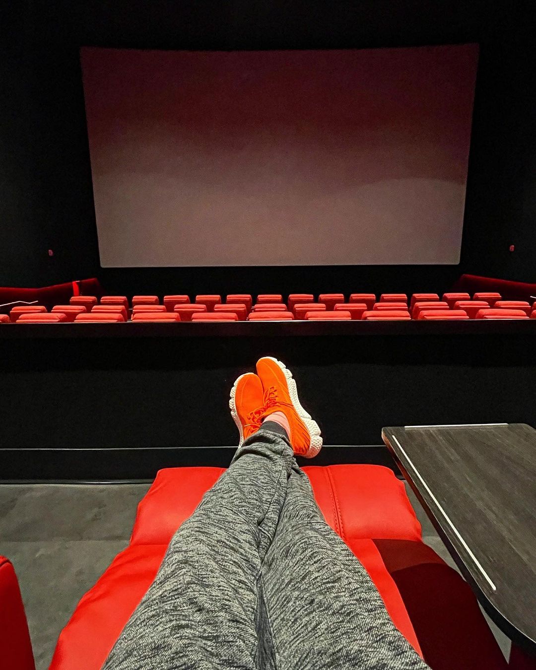A person extends their crossed legs on a red recliner in front of an empty theater with a blank screen.