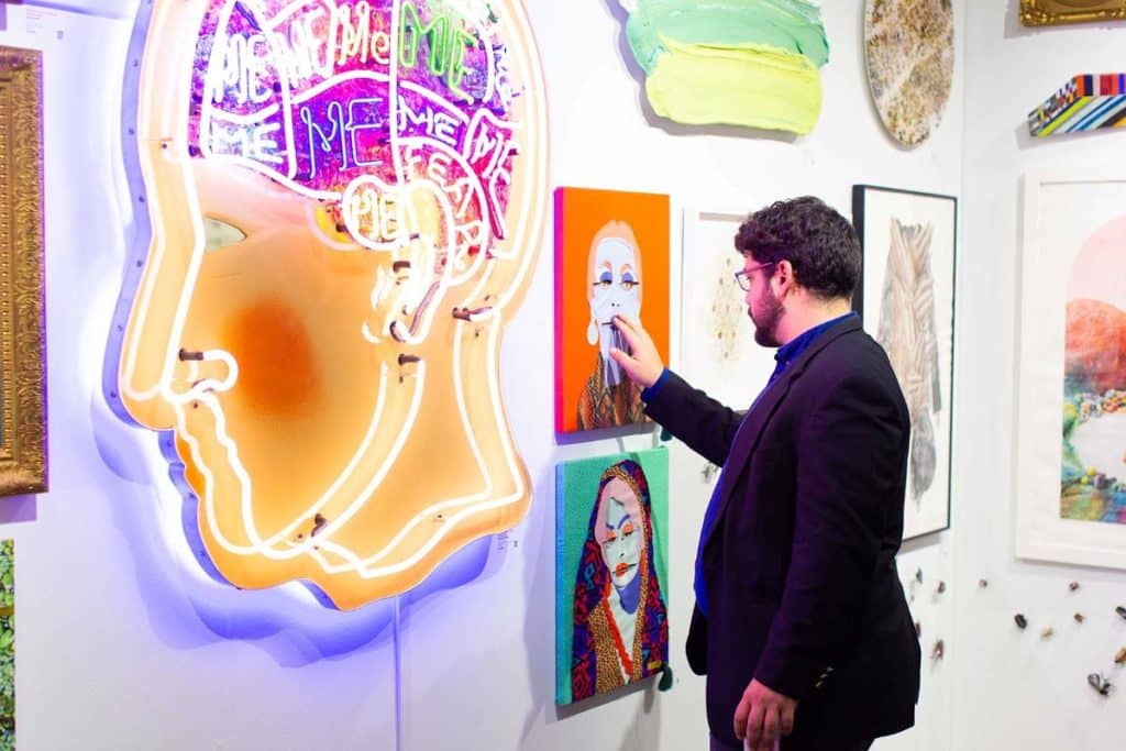 A man looks at colorful art pieces including a large neon profile head.