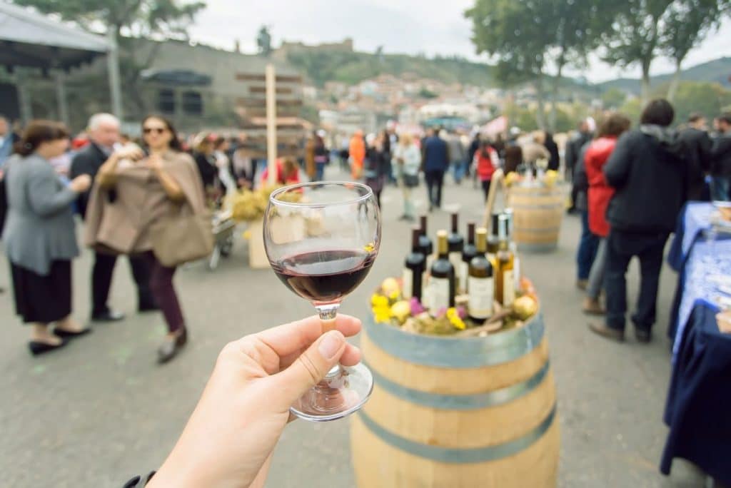 A person holds a glass of wine out in front of the camera with a table of wine bottles and crowds of people in the background.