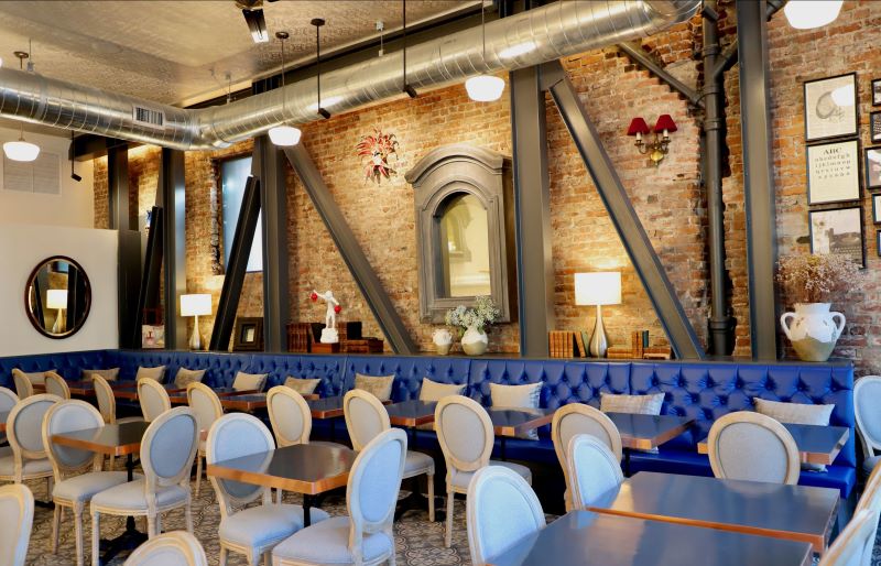 An empty dining room with a blue plush sofa and pale blue chairs beneath a brick wall and industrial elements.
