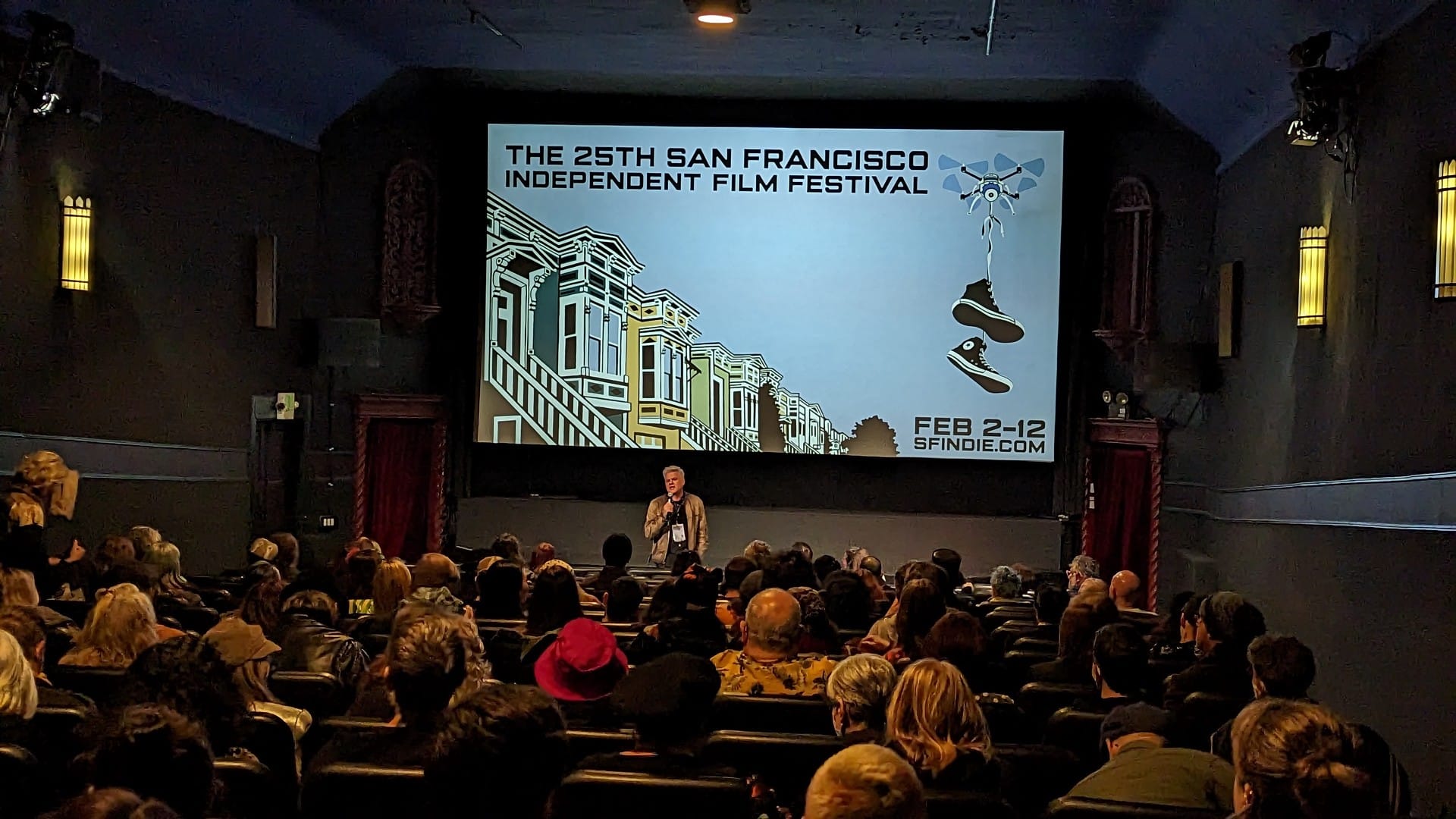 A man with a microphone speaks to an audience in front of a screen reading "25th San Francisco Independent Film Festival" 