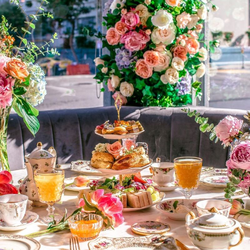 A table set with a tiered platter of pastries, sandwiches, and tea surrounded with colorful flower decorations.