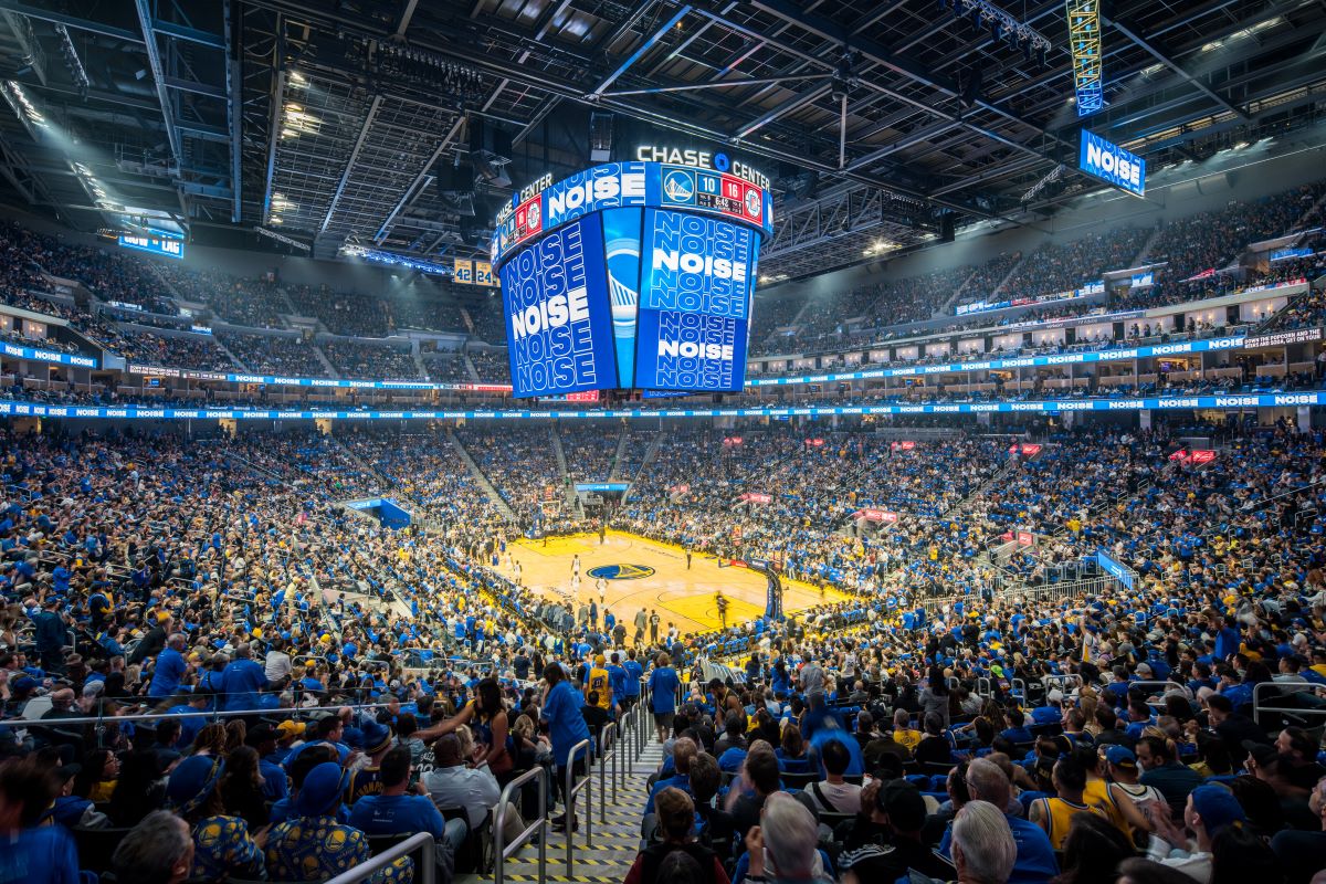Wide shot of Chase Center arena interior showing the scorehead above a brightly lit basketball court surrounded by thousands of fans.