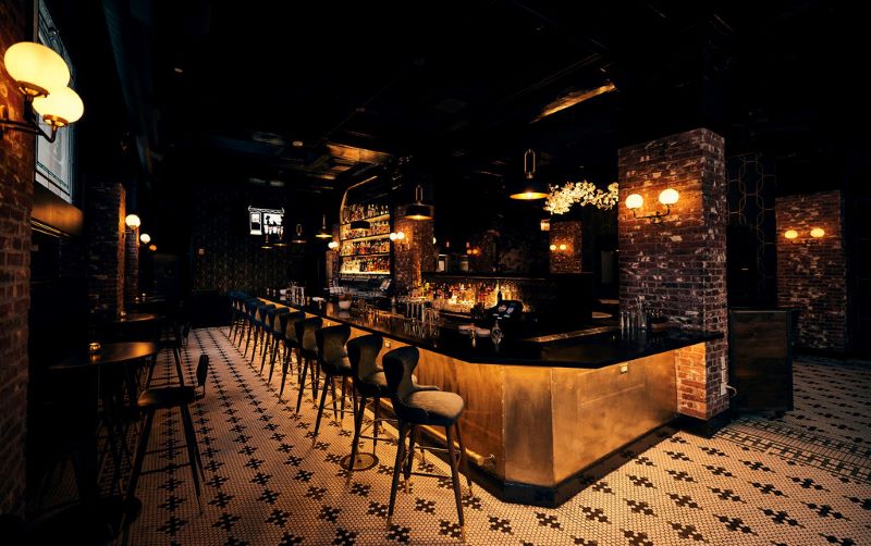 Bar seating at the Harlequin with black and white patterned tile, dark ceiling and walls, and gold lights.