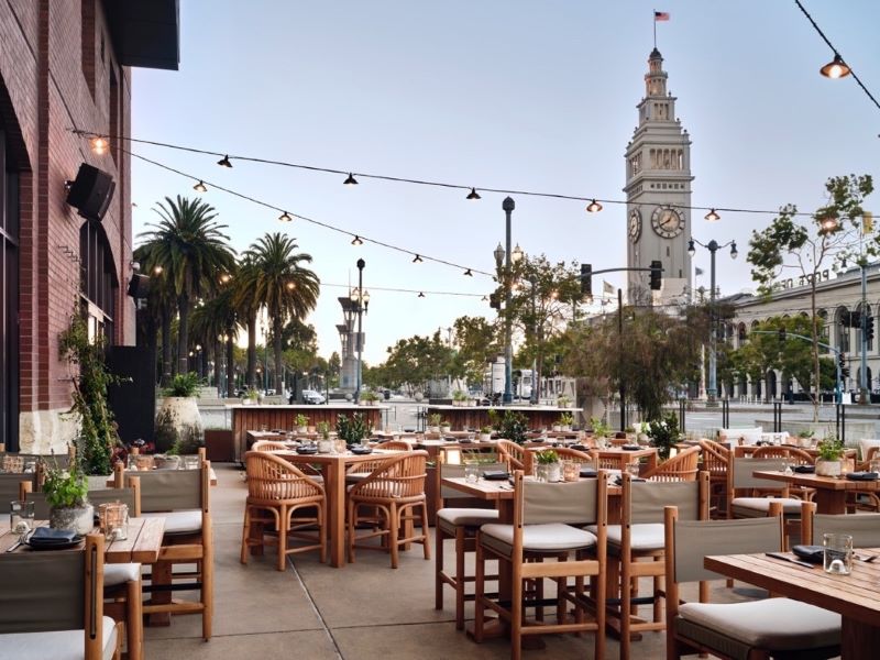 Outdoor dining area at 1 Hotel SF with string lights and the Ferry Building in the background.