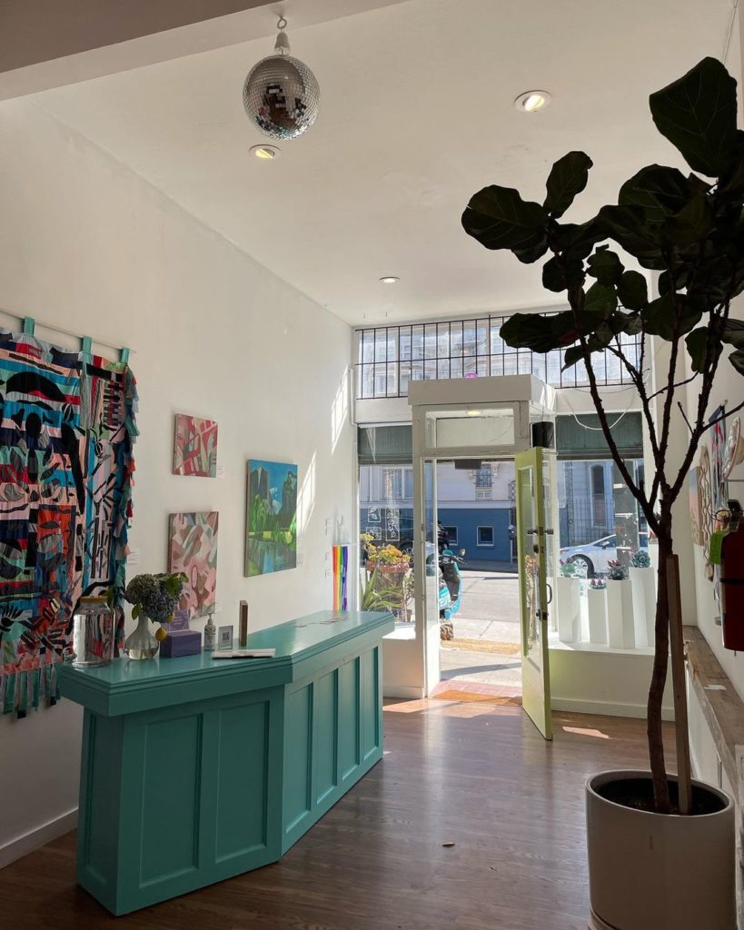 Inside of a gallery with a blue counter, large plant, and a door open to the street.