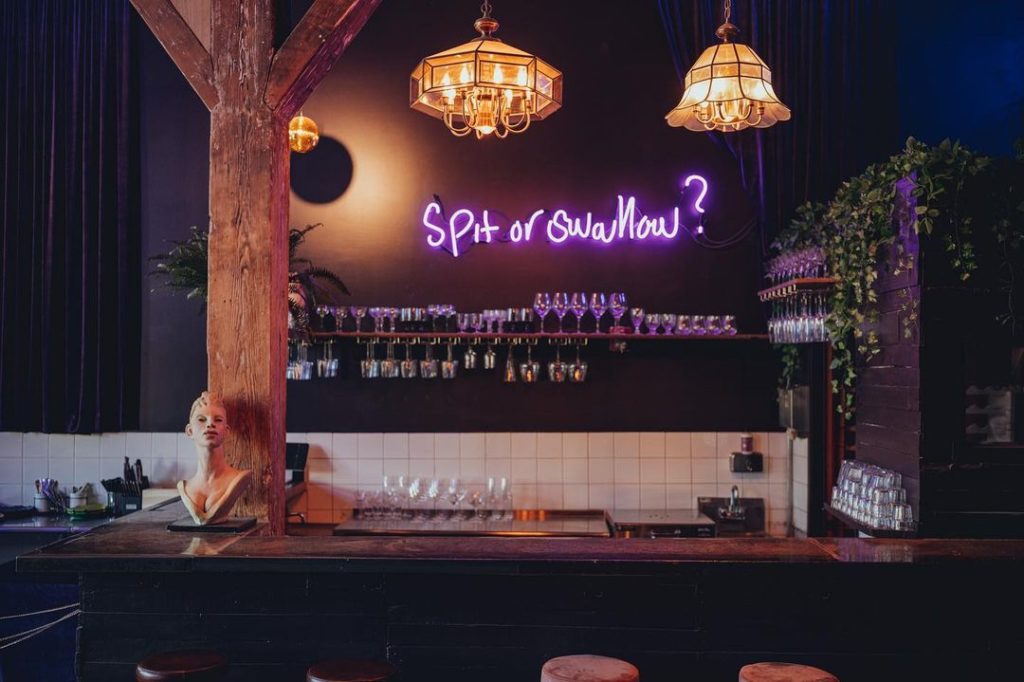 A bar with dark walls bottles of wine on the wall with a neon sign that reads "spit or swallow?"
