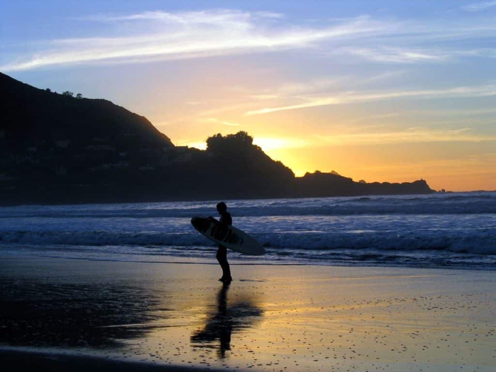 A person holding a surf board walking on the beach at sunset