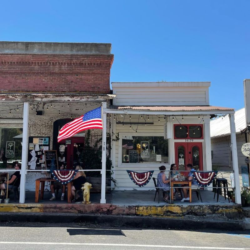 People eat at outdoor tables in front of historic buildings decorated in American flags.