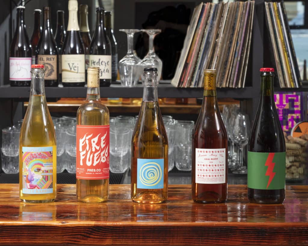 Five wine bottles on a bar with records and more bottles in the background.