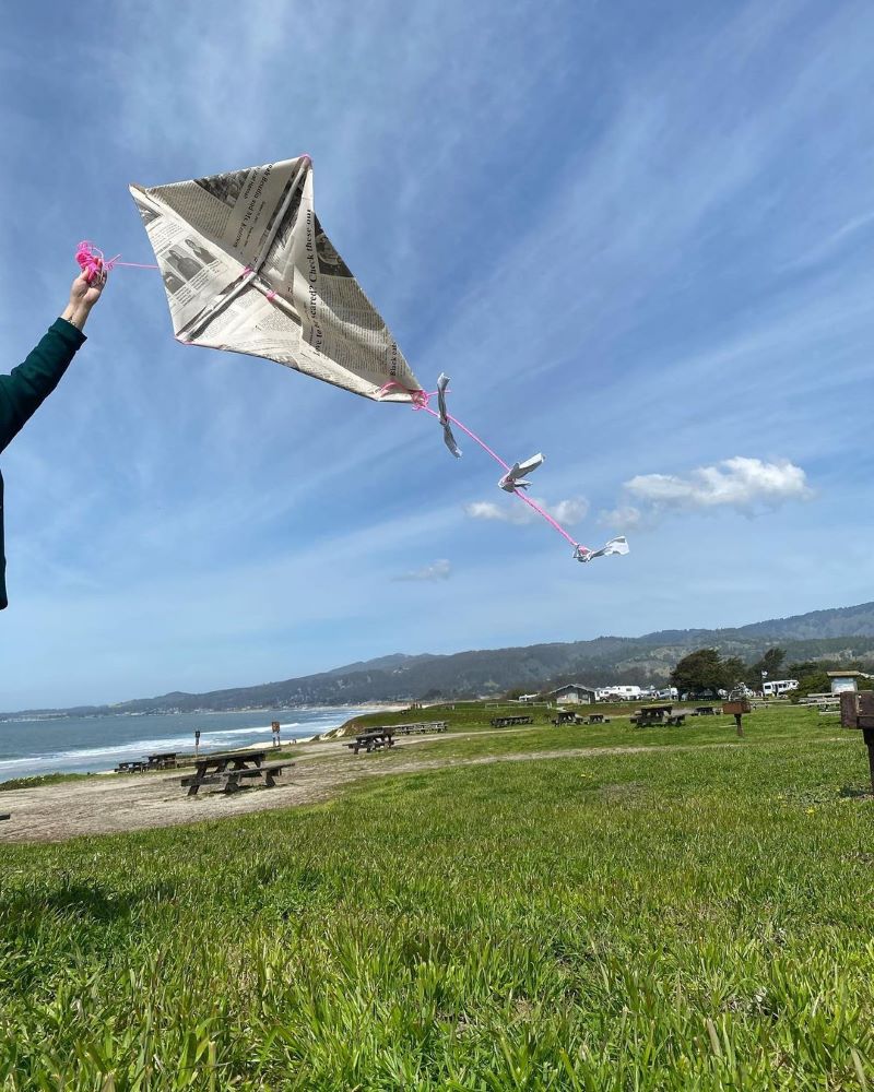 A person flies a kite made with newspaper on a grassy field near the beach.