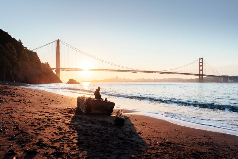 A person sits on a log on the beach with the Golden Gate Bridge in the background. 