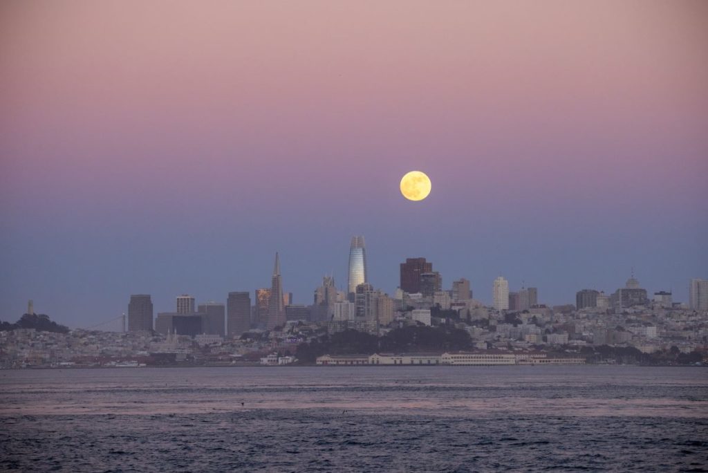 A full moon rises over the SF skyline at sunset.